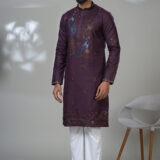 Man wearing a plum-colored embroidered kurta with blue peacock motifs, white pants, and brown formal loafers, standing beside a modern white chair
