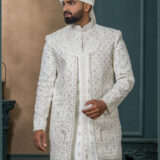 Groom in an ivory sherwani with geometric and floral zari embroidery, styled with a white turban adorned with feather and jewel embellishment.
