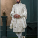 Groom in an ivory sherwani with geometric and floral zari embroidery, styled with a white turban adorned with feather and jewel embellishment.