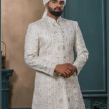 Ivory mirror-work sherwani with intricate embroidery, styled with a white turban and feather for a royal groom look.