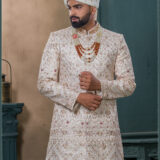 Champagne gold sherwani with red and gold embroidery, paired with layered pearl and stone mala and an ivory turban with feather embellishment.