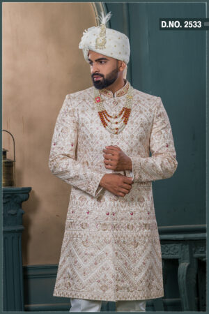 Champagne gold sherwani with red and gold embroidery, paired with layered pearl and stone mala and an ivory turban with feather embellishment.