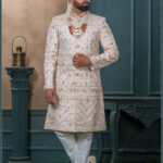 Champagne gold sherwani with red and gold embroidery, paired with layered pearl and stone mala and an ivory turban with feather embellishment.