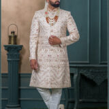 Champagne gold sherwani with red and gold embroidery, paired with layered pearl and stone mala and an ivory turban with feather embellishment.