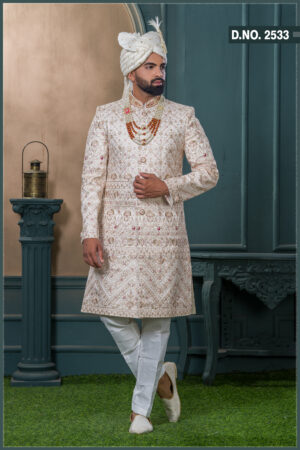 Champagne gold sherwani with red and gold embroidery, paired with layered pearl and stone mala and an ivory turban with feather embellishment.