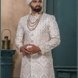 Ivory gold sherwani with detailed embroidery, red collar highlights, layered pearl and ruby mala, and ivory turban with feather accent.