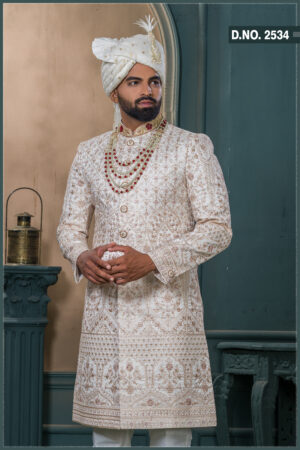 Ivory gold sherwani with detailed embroidery, red collar highlights, layered pearl and ruby mala, and ivory turban with feather accent.