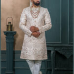 Ivory gold sherwani with detailed embroidery, red collar highlights, layered pearl and ruby mala, and ivory turban with feather accent.