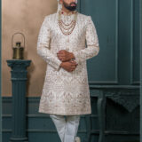 Ivory gold sherwani with detailed embroidery, red collar highlights, layered pearl and ruby mala, and ivory turban with feather accent.