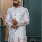 White sherwani with golden zari and ruby red embroidery, detailed with red buttons and paired with an ivory turban adorned with feather and gold brooch.