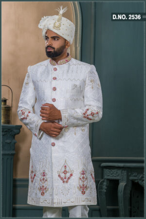 White sherwani with golden zari and ruby red embroidery, detailed with red buttons and paired with an ivory turban adorned with feather and gold brooch.