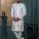 White sherwani with golden zari and ruby red embroidery, detailed with red buttons and paired with an ivory turban adorned with feather and gold brooch.