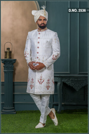 White sherwani with golden zari and ruby red embroidery, detailed with red buttons and paired with an ivory turban adorned with feather and gold brooch.